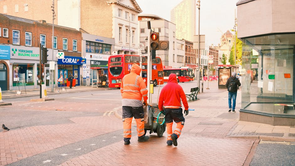 Two waste collection workers dressed in high-visibility orange and reflective striped trousers and jackets are pushing a wheeled trash container along a city sidewalk near a busy intersection. The wor