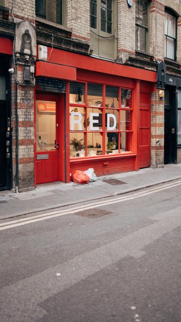 Two waste collection workers dressed in high-visibility orange and reflective striped trousers and jackets are pushing a wheeled trash container along a city sidewalk near a busy intersection. The wor